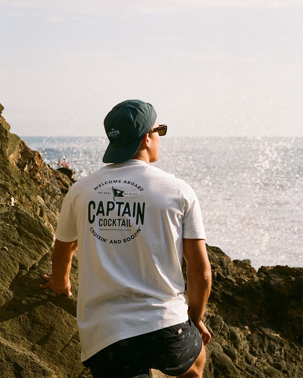 Men's white graphic t-shirt featuring 'Captain Cocktail' text and a martini glass, wearing the shirt on rocks near the ocean.