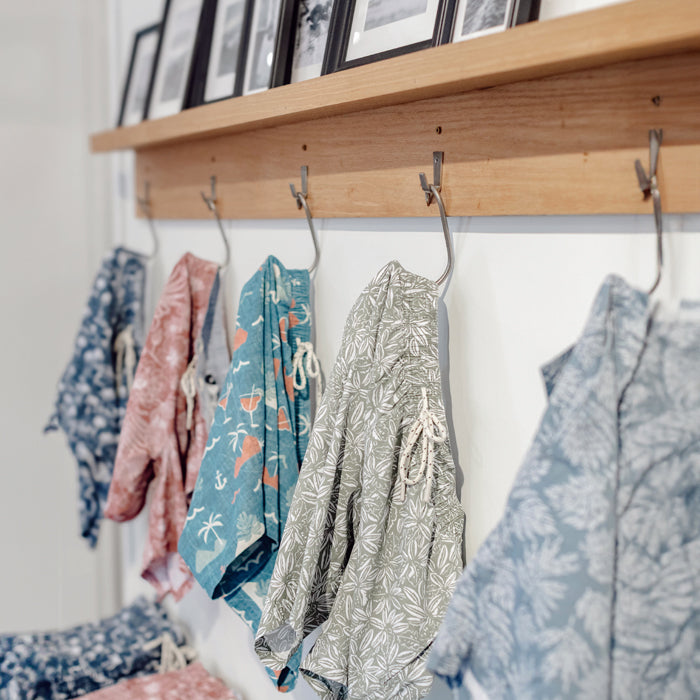An interior wall decorated with framed surf-themed artwork, including black and white photos of surfers, and two decorative pennants: one green with 'CALIFORNIA' and one blue with 'HUNTINGTON BEACH', with a green potted plant in the foreground, reflecting the coastal style of Toes on the Nose in Huntington Beach