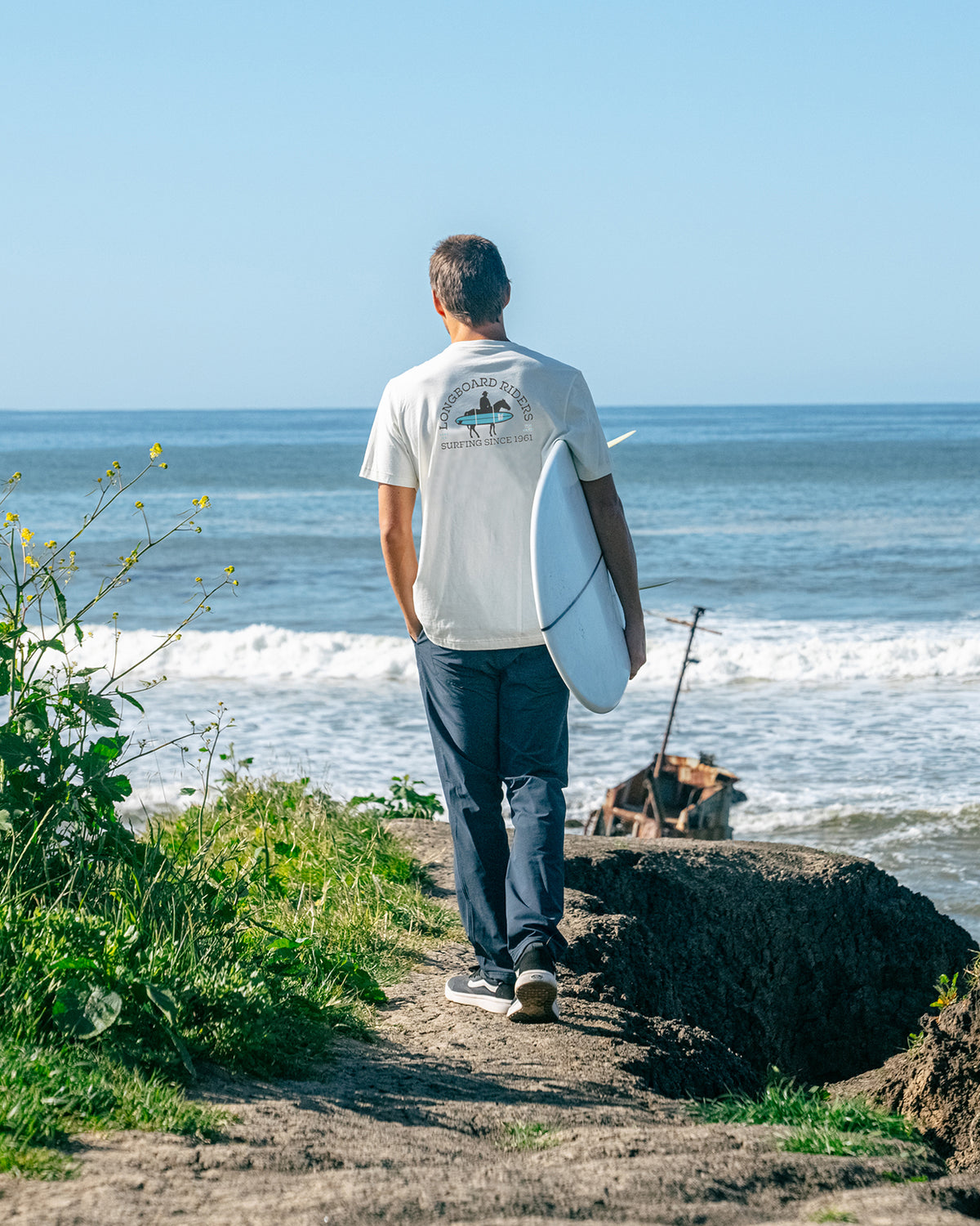 Men's white t-shirt with "LONGBOARD RIDERS" and a surfboard graphic, worn by a man carrying a surfboard on the beach.