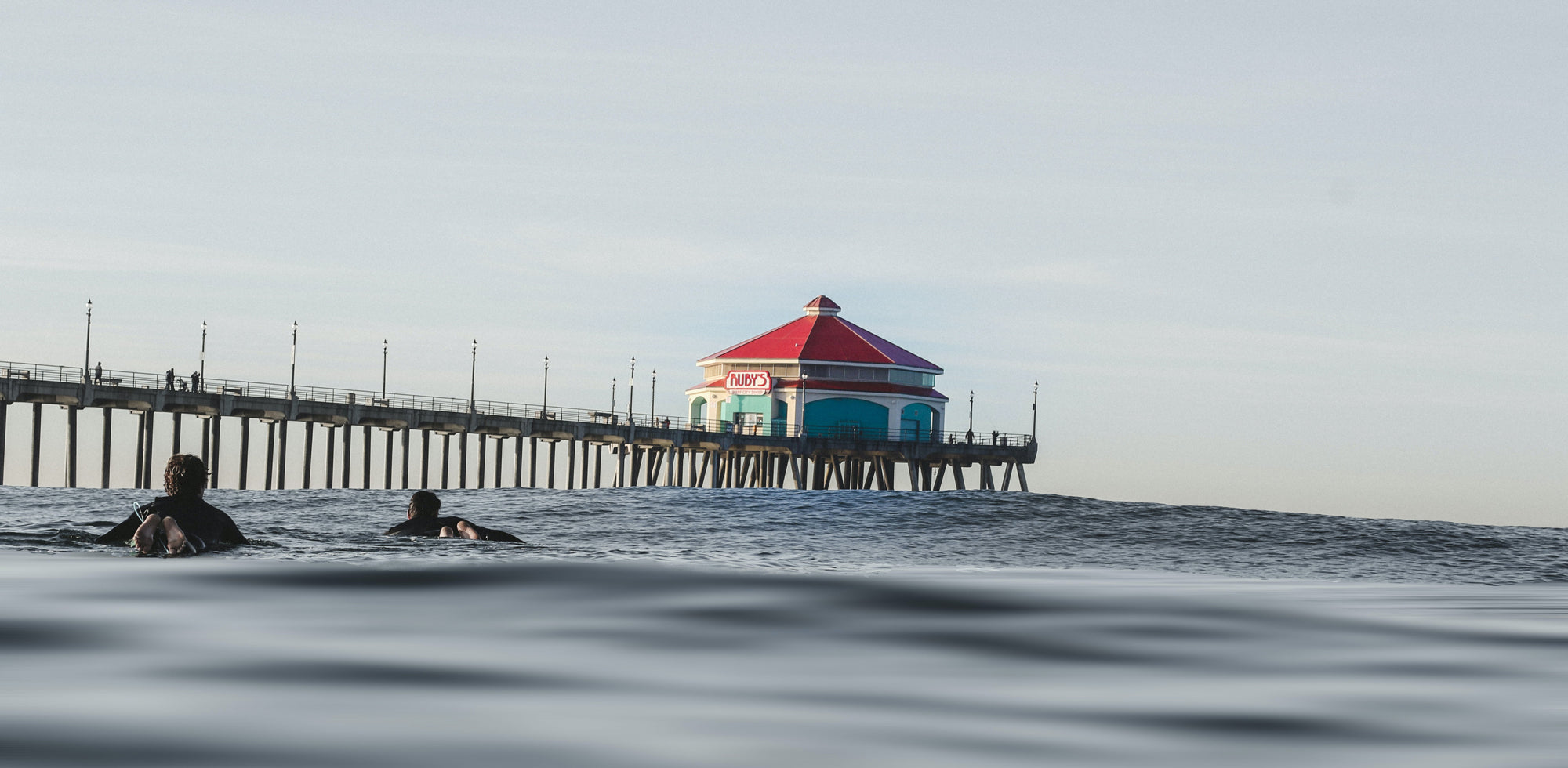 Surfers in the ocean near Huntington Beach Pier with Ruby’s Diner in the background, reflecting the relaxed surf lifestyle and coastal vibe of Toes on the Nose apparel