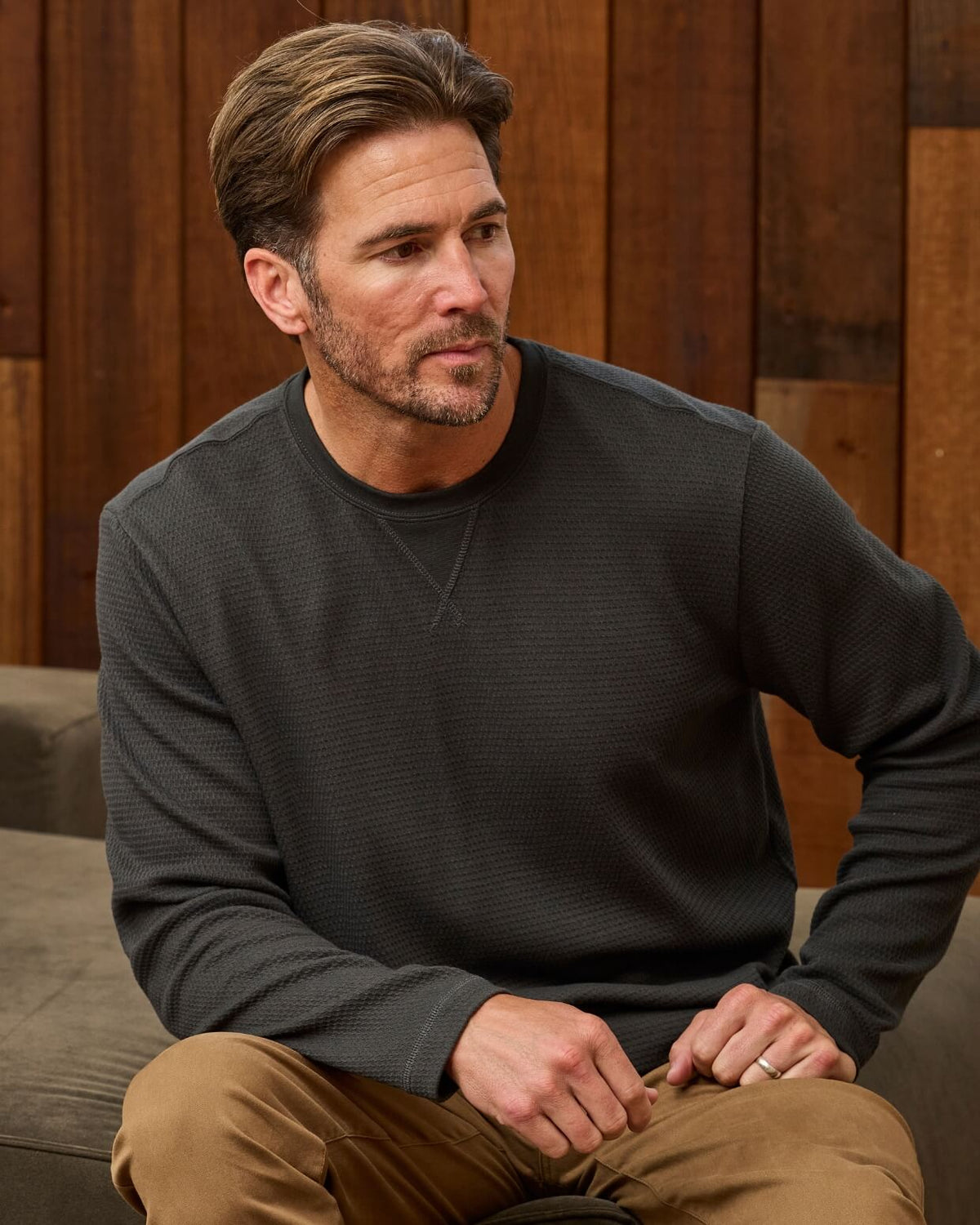 A man wearing a dark charcoal gray ribbed long-sleeve crewneck sweater and tan pants, sitting in a relaxed pose against a warm wood-paneled background. #color_faded black