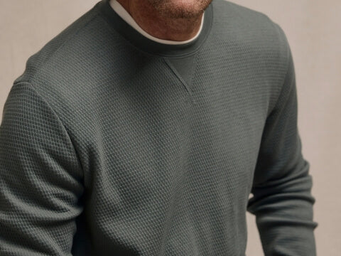 A close-up of a man wearing a textured, long-sleeve crewneck sweater in a muted sage green color, layered over a white undershirt.