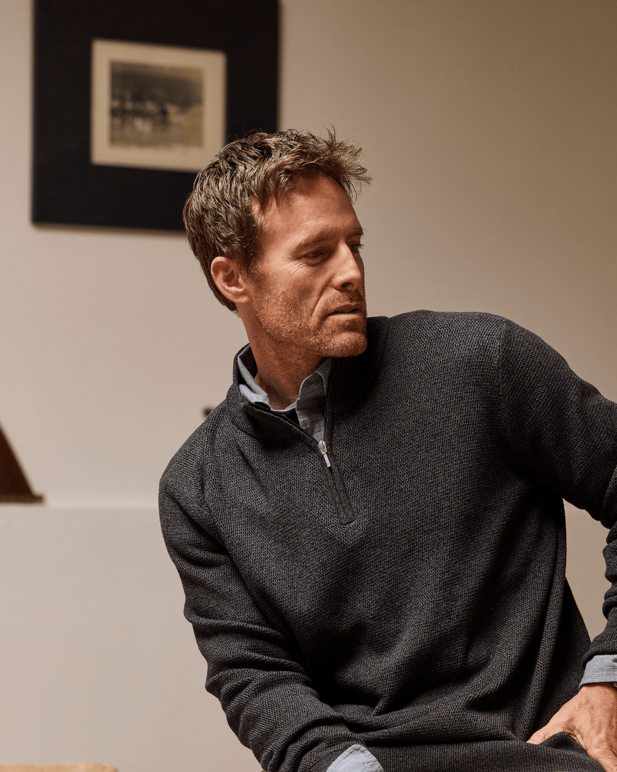 A man wearing a dark gray quarter-zip sweater over a light blue collared shirt, seated indoors against a beige wall with a framed picture in the background. #color_easton black