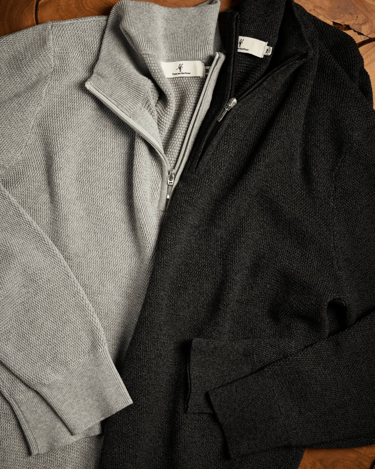 Two quarter-zip knit sweaters laid flat on a wooden surface: one light gray and one black. #color_easton grey