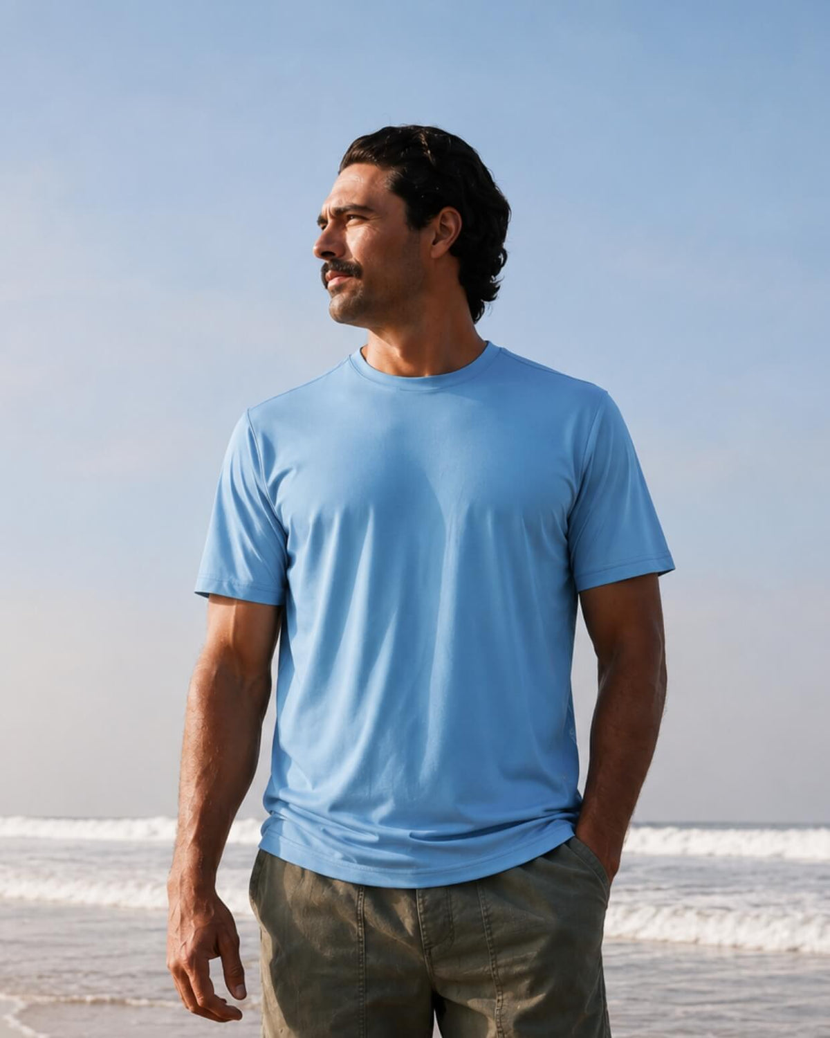 A man wearing a light blue crew-neck t-shirt and olive green pants standing on a beach with the ocean and a clear blue sky in the background. #color_ocean crew