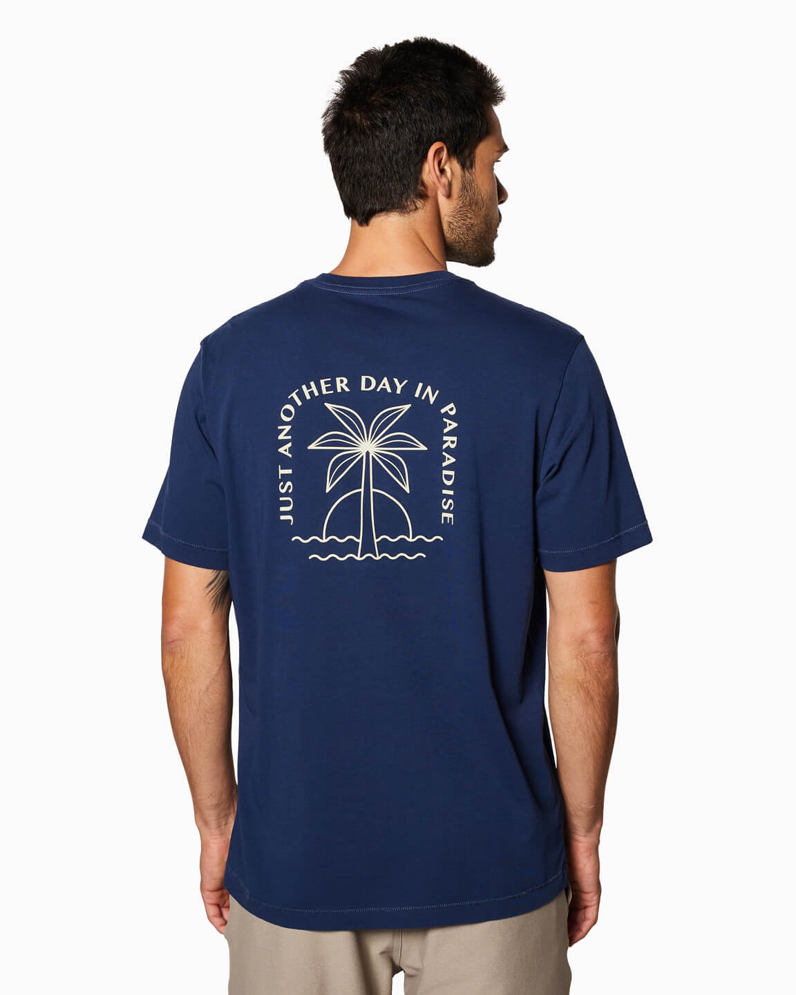 Men's navy t-shirt with stylized palm tree and "Just Another Day in Paradise" text, worn by a model standing indoors.