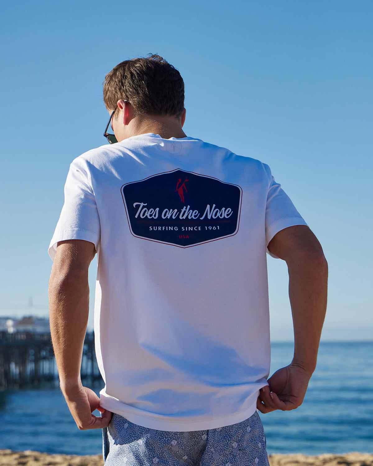 Men's white t-shirt featuring a large back print with the Toes on the Nose logo and 'Surfing Since 1961', worn by a model with a scenic beach background.