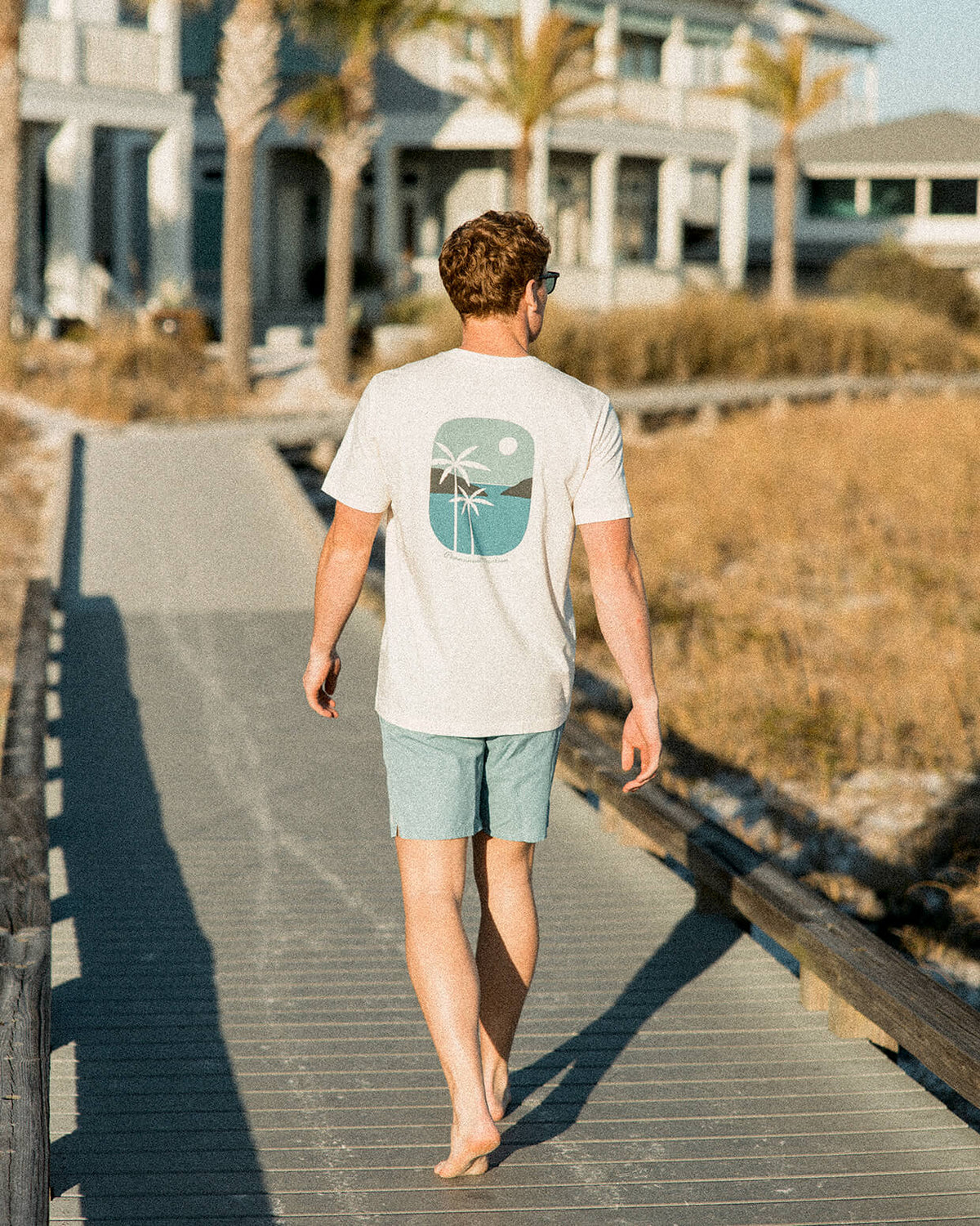 Men's off-white t-shirt featuring a retro beach graphic with palm trees, walking on a boardwalk near the beach.