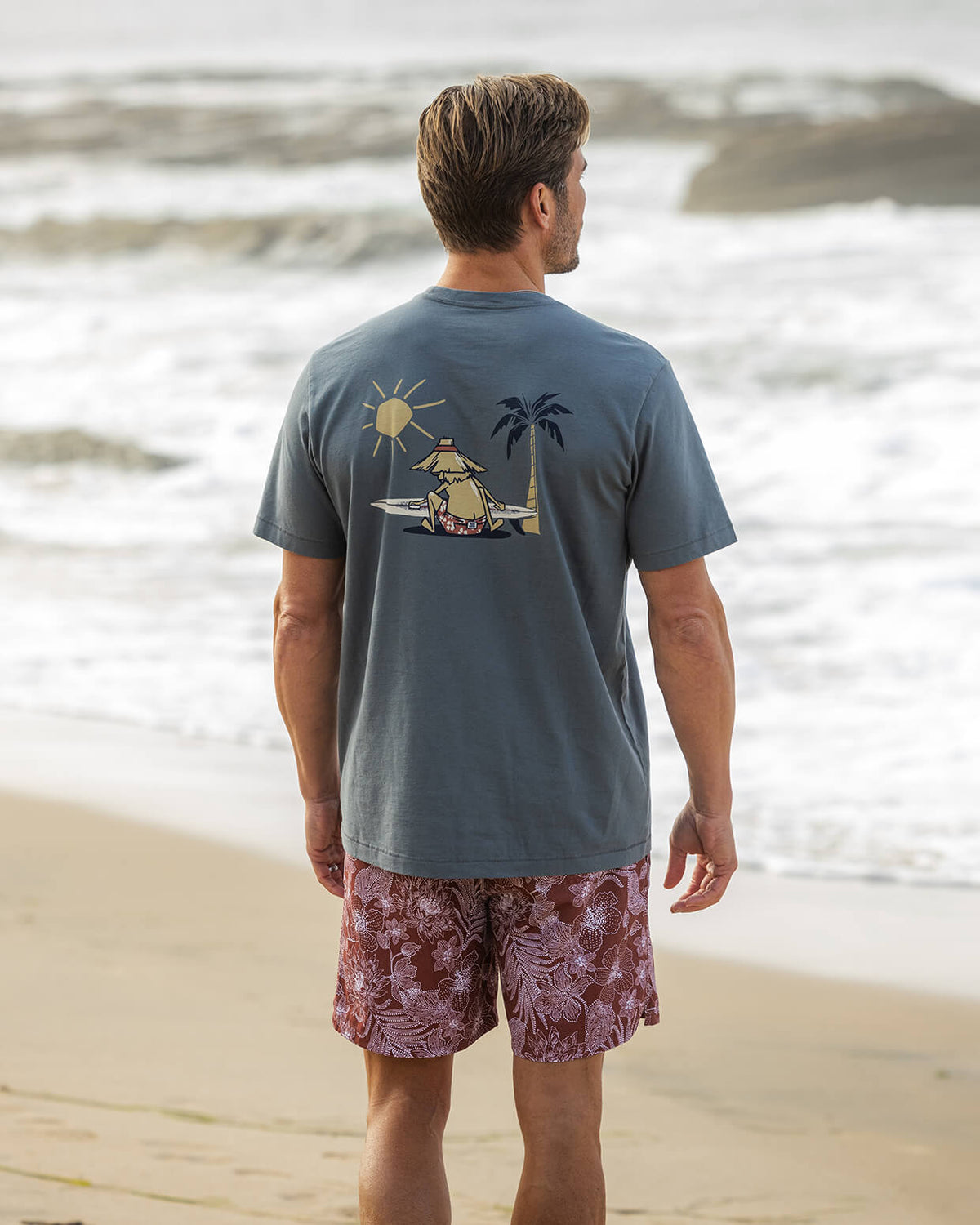 Men's blue graphic t-shirt featuring a tropical island scene with a surfer, angled on a beach background.