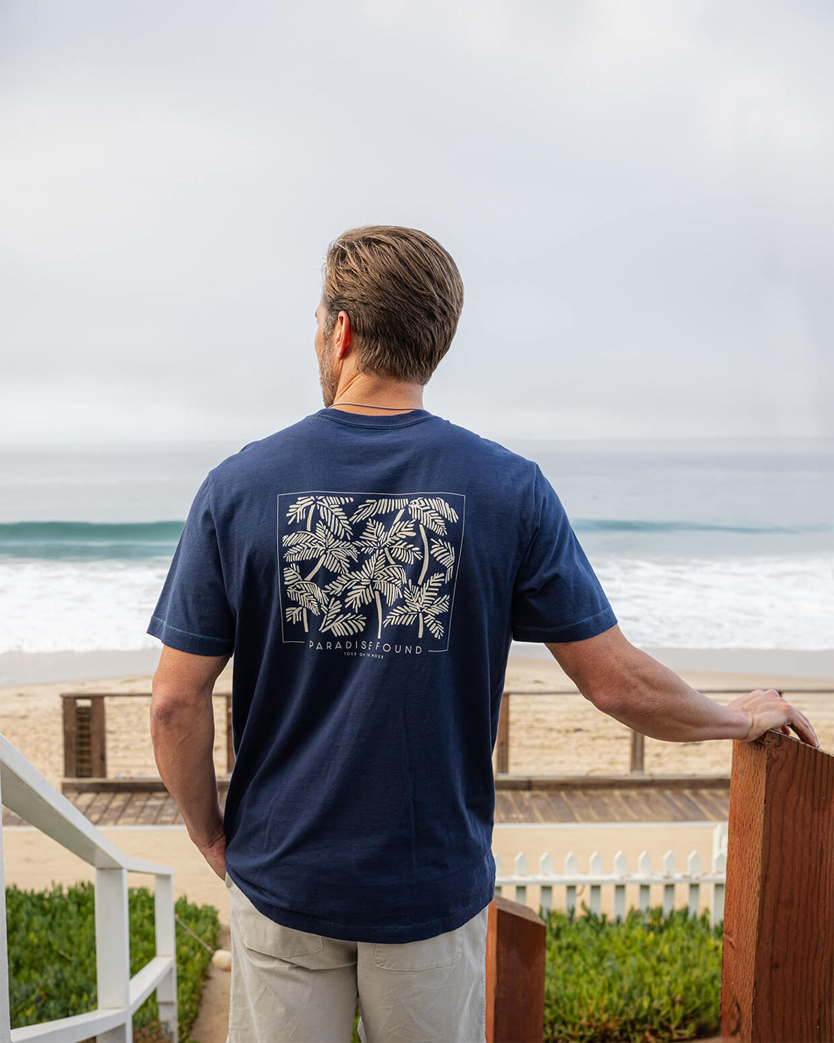 Men's navy t-shirt with palm leaf design and the "PARADISE FOUND" logo, worn by a man at the beach with the ocean in the background.