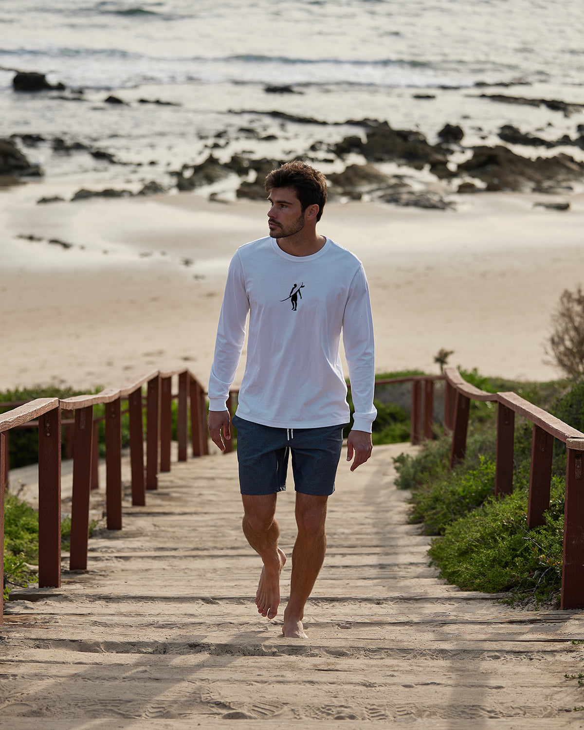 Men's white long-sleeve t-shirt featuring a surfer graphic on the chest, walking barefoot on the beach.