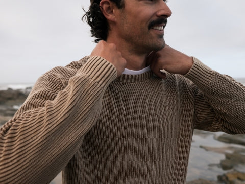 A man wearing a brown ribbed knit crewneck sweater over a white undershirt, standing outdoors near a rocky shoreline under an overcast sky.