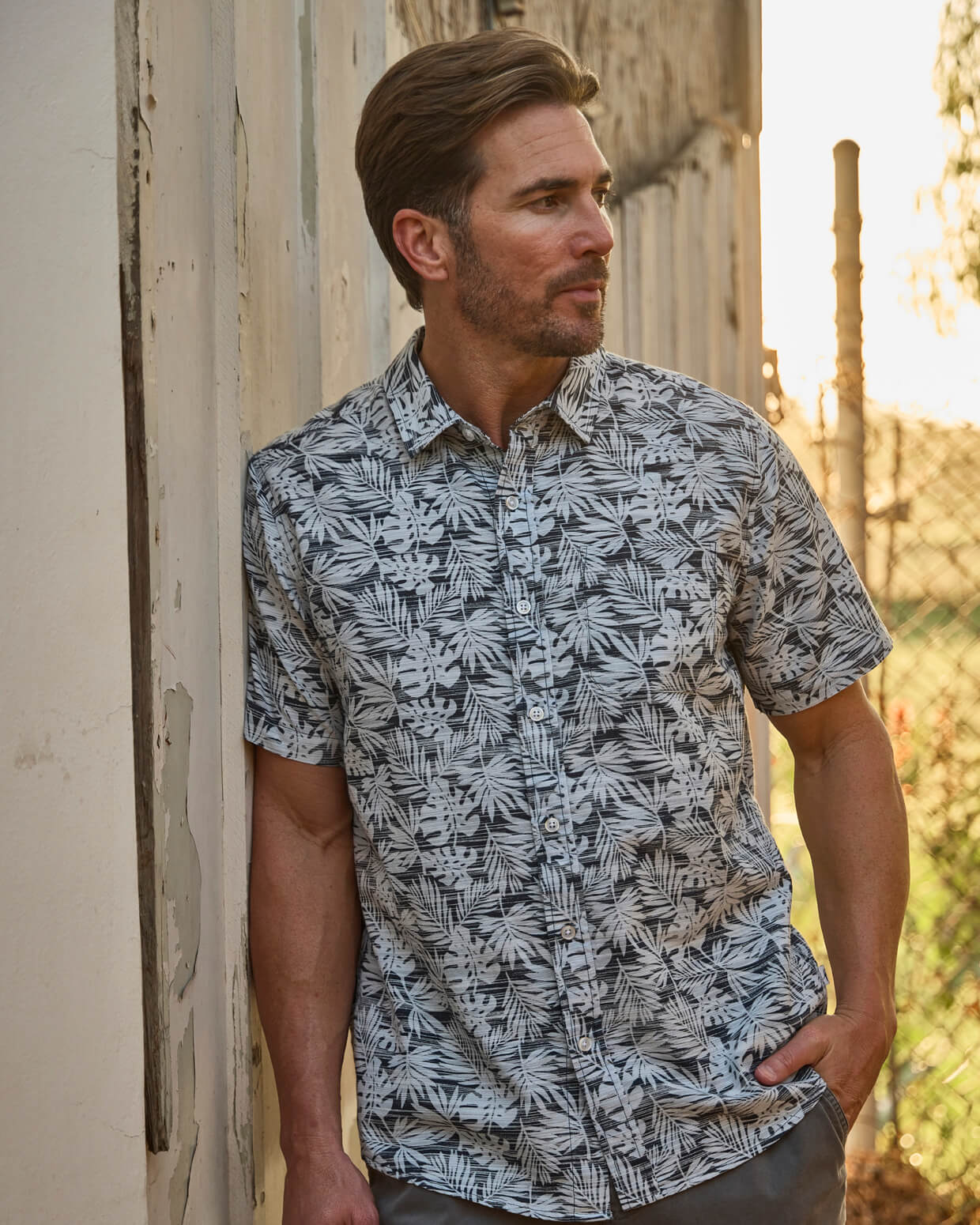 Man wearing a patterned shirt standing against a rustic wall with a natural background