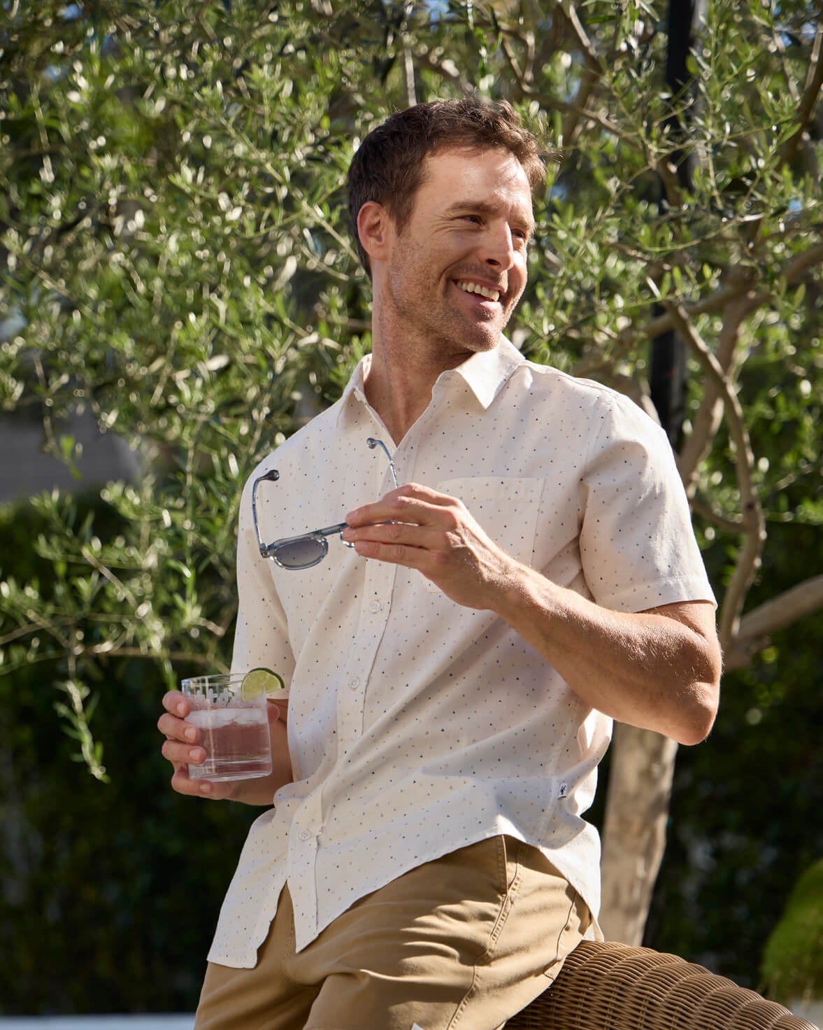 Man in a light shirt and khaki pants holding a glass outdoors with greenery in the background #color_washed white naples