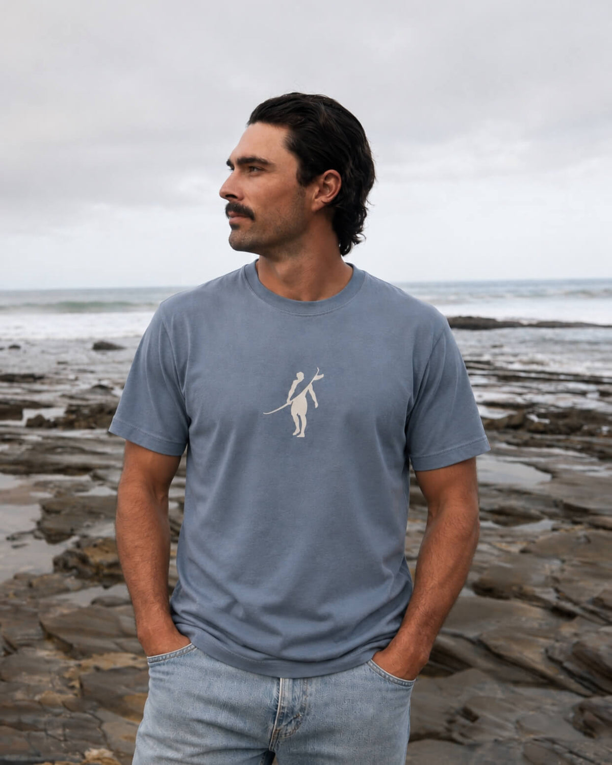 A man wearing a slate blue crewneck t-shirt with a white minimalist graphic of a surfer, standing on a rocky beach under an overcast sky. #color_blue flint