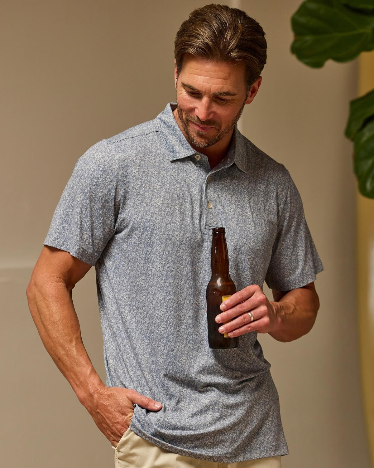 A man wearing a textured heather gray short-sleeve polo shirt and khaki pants, holding a brown glass beer bottle against a warm beige background. #color_grove blue