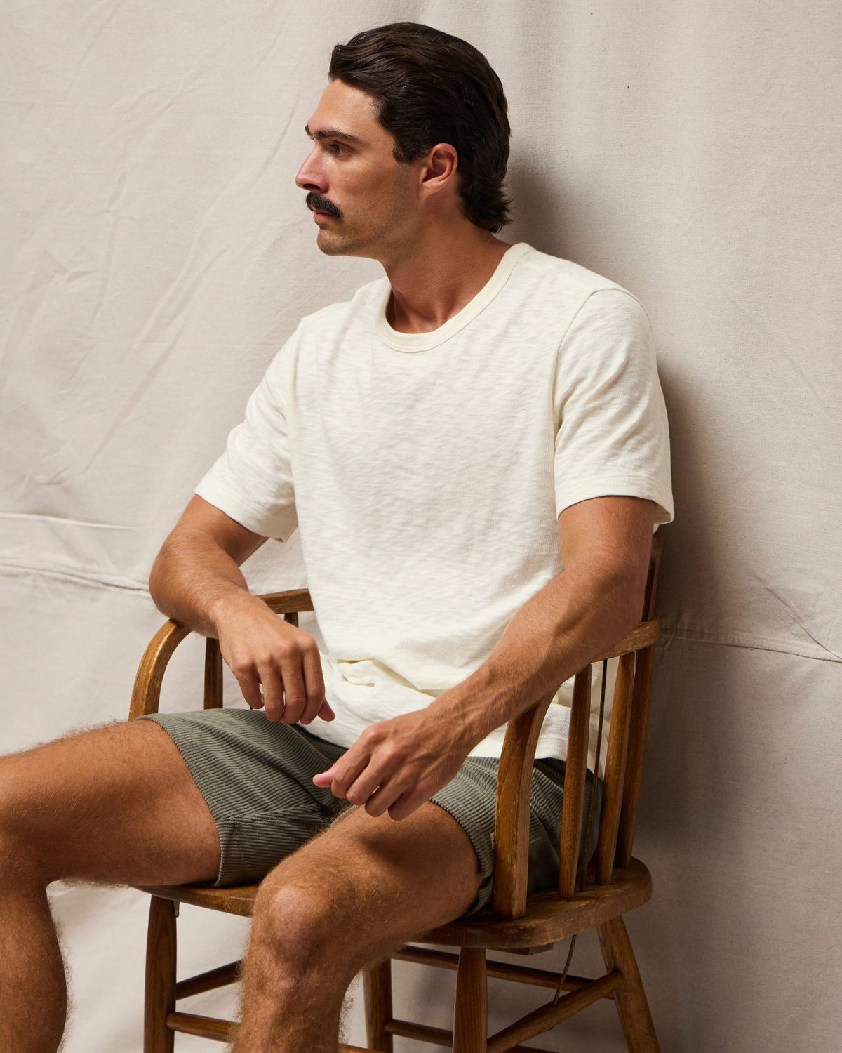 A man with a mustache sits on a wooden chair, wearing a light cream-colored short-sleeve t-shirt and olive green shorts against a neutral draped fabric backdrop. #color_natural slub