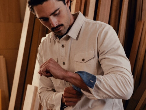 A man wearing a light beige long-sleeve button-up shirt with a chest pocket, shown adjusting his cuff in a warm, wood-paneled setting.