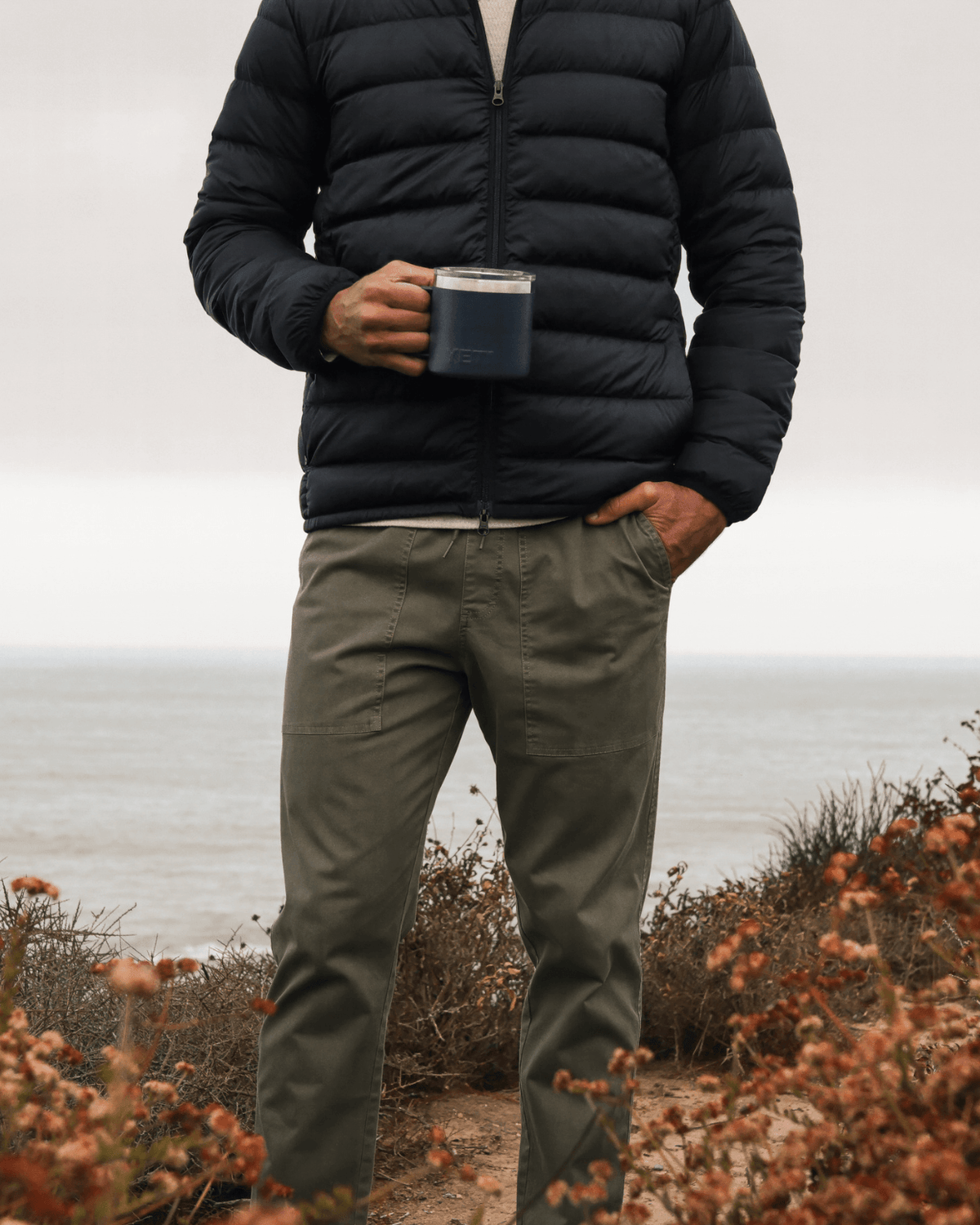 Person wearing a black puffer jacket and green pants holding a mug outdoors with a neutral background #color_alston olive