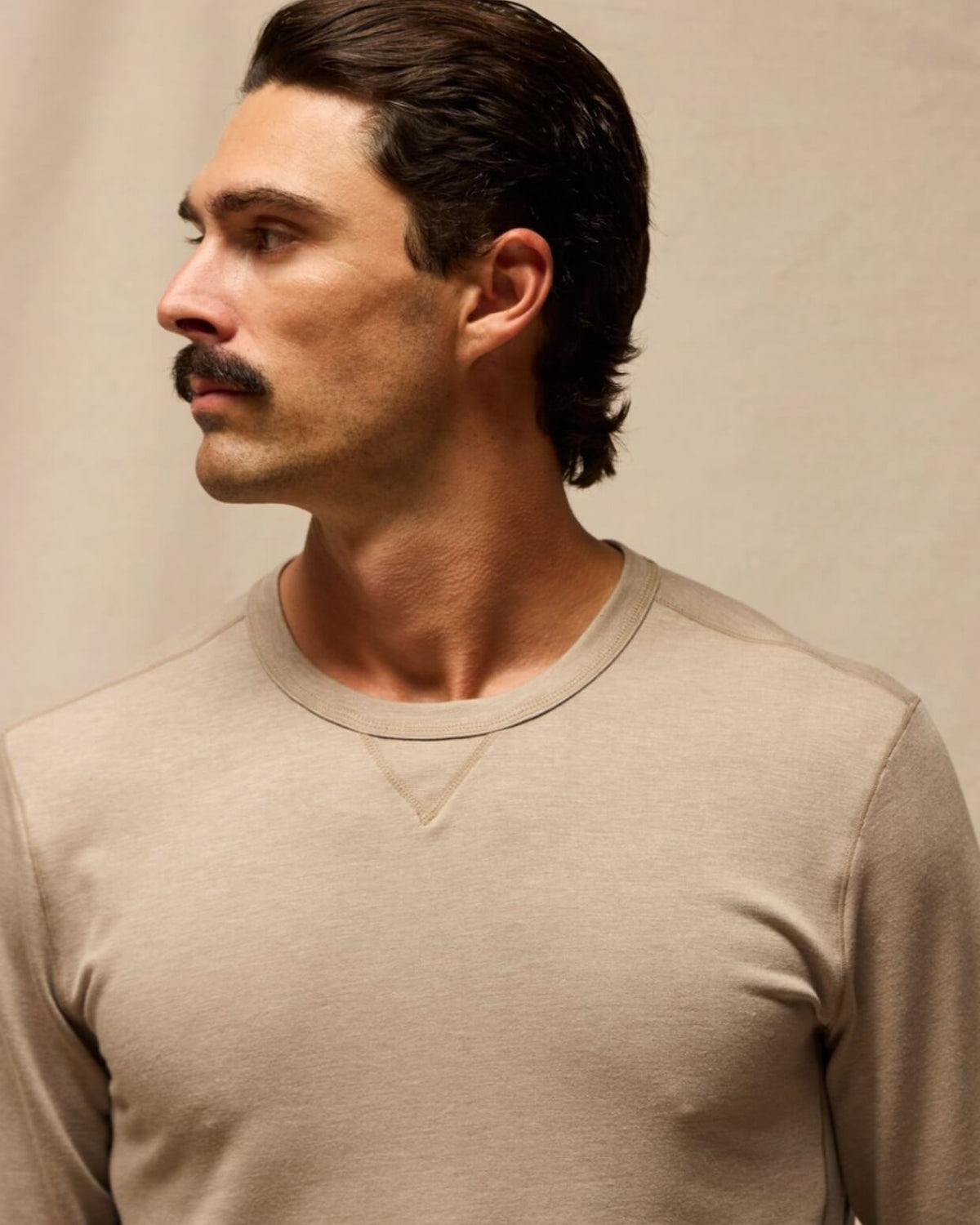 A man with a mustache wearing a light beige crewneck long-sleeve shirt, shown in a close-up profile against a neutral beige background. #color_vintage khaki