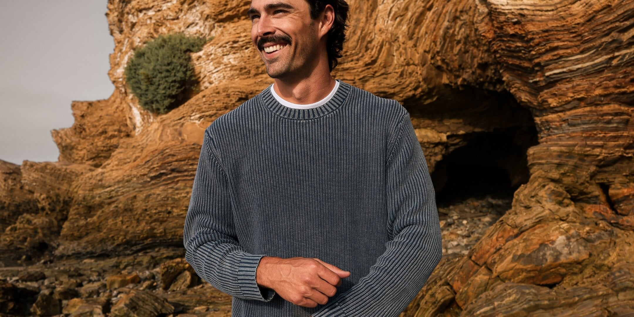 A man with a mustache wearing a dark gray ribbed knit sweater, standing outdoors in front of a large brown rock formation.