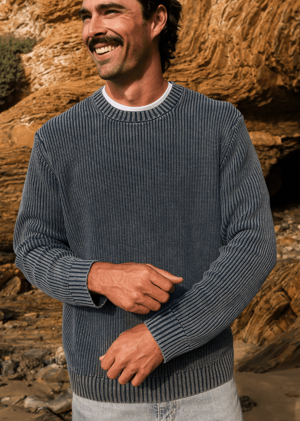 A man wearing a dark gray ribbed knit crewneck sweater, standing outdoors in front of a rocky, sandy desert landscape.