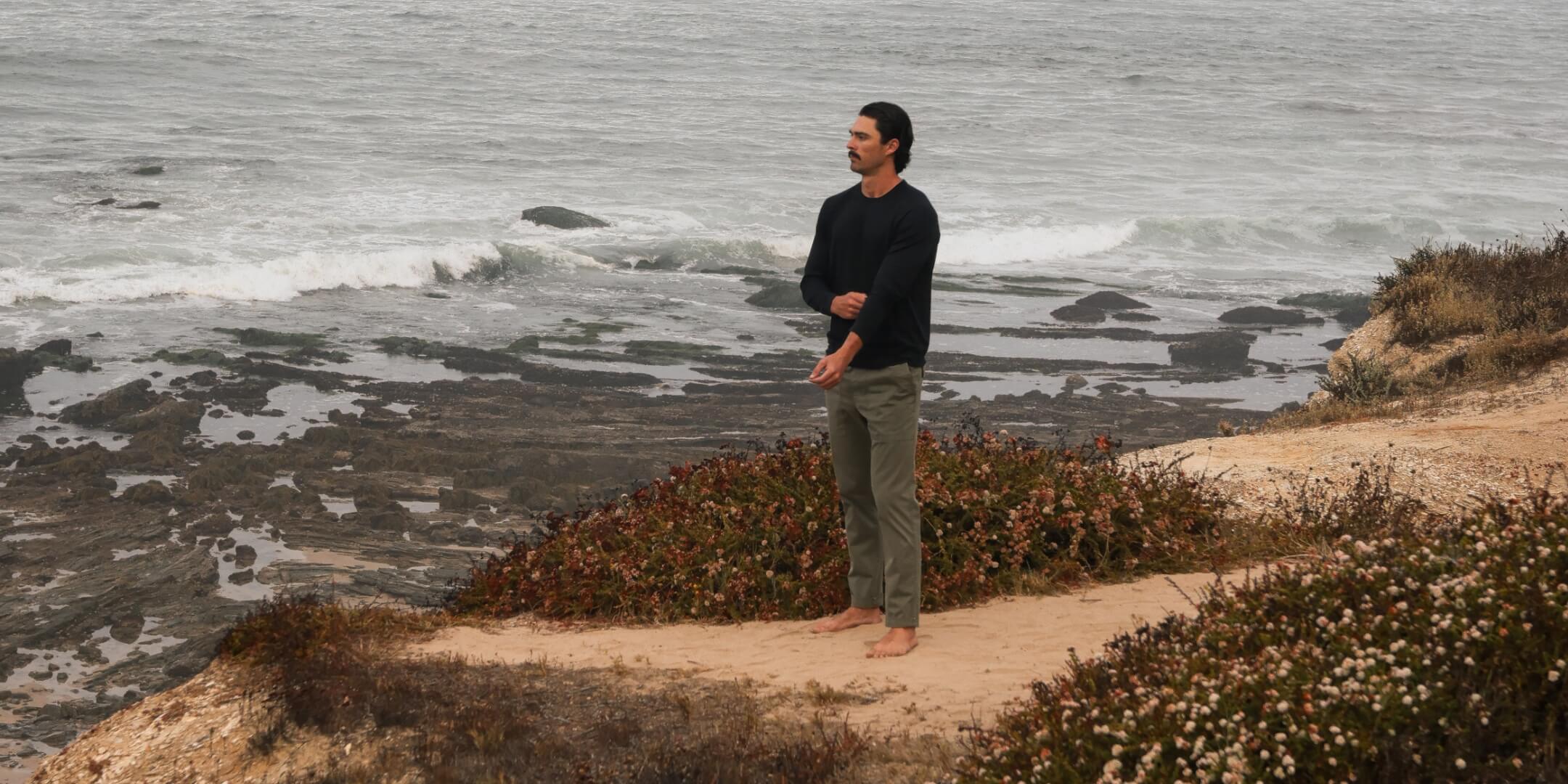A man stands barefoot on a sandy coastal path overlooking the ocean, wearing a dark long-sleeve shirt and olive green pants.