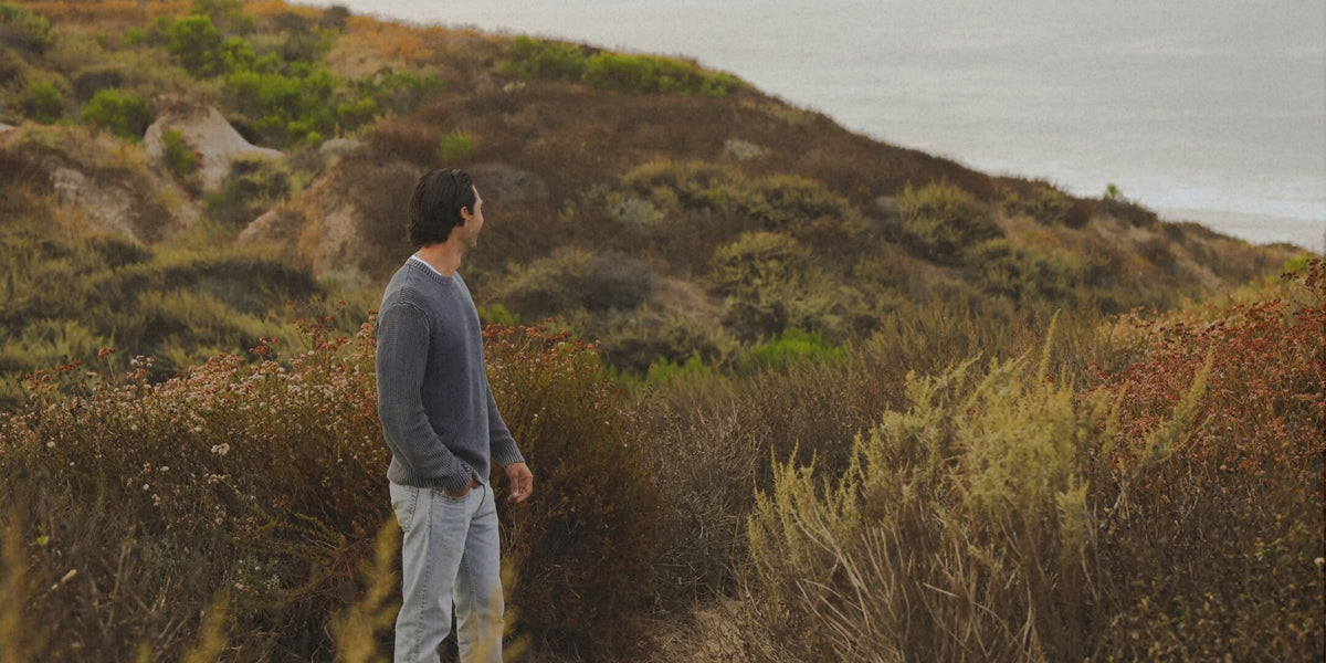 A man in a gray long-sleeve shirt and light gray jeans stands on a grassy hillside overlooking a coastal ocean landscape.