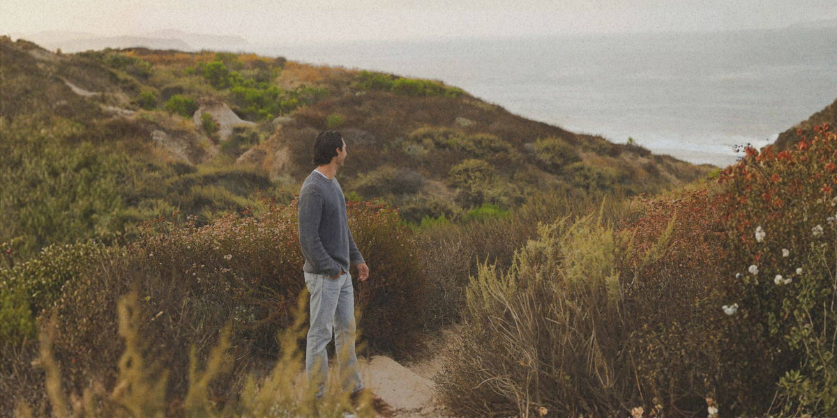A man in a gray long-sleeve shirt and light gray jeans stands on a dirt path in a coastal shrub-covered landscape, looking out toward the ocean under an overcast sky.