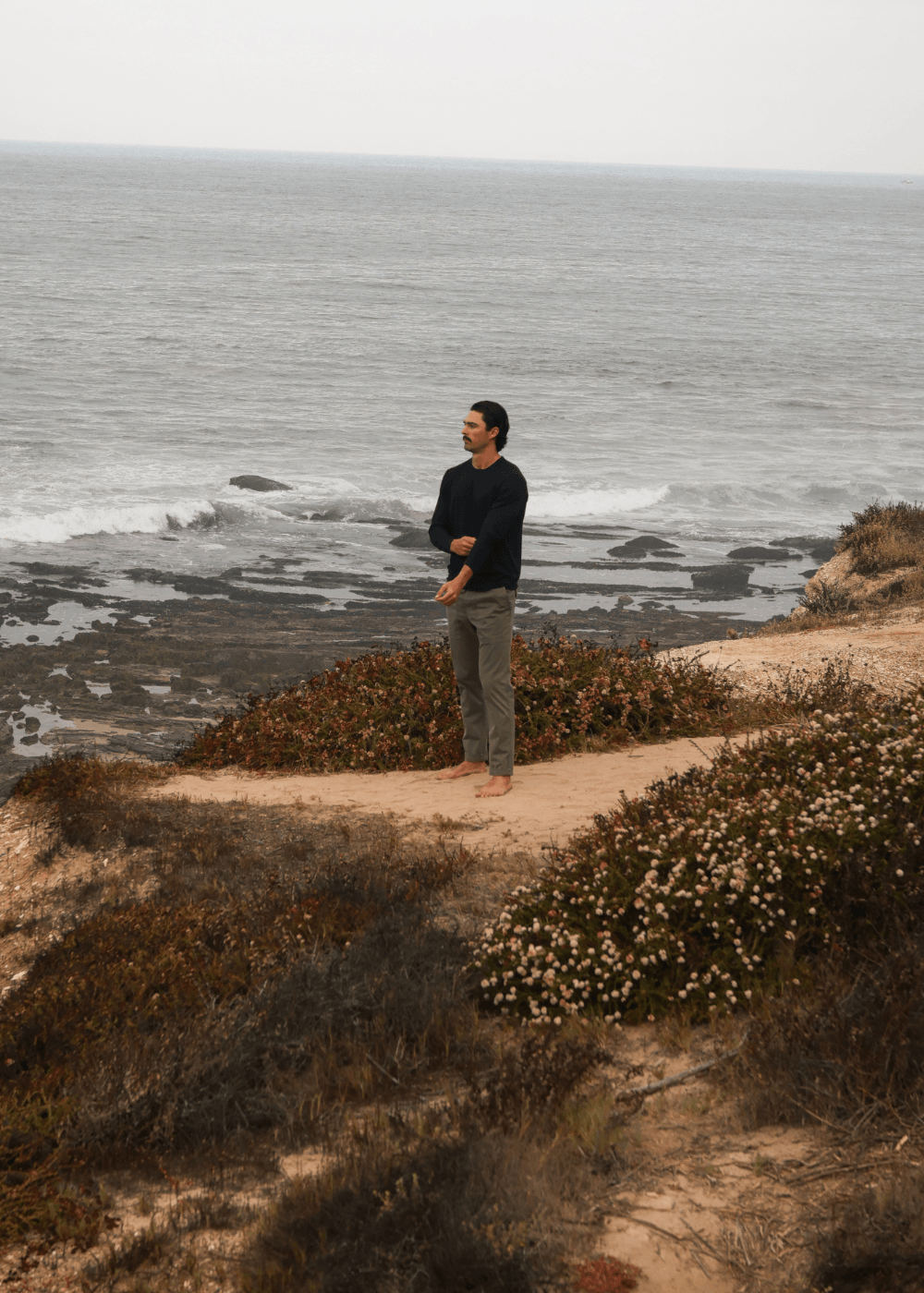 A man stands barefoot on a sandy path overlooking the ocean, wearing a dark long-sleeve shirt and gray pants, with coastal shrubs and rocks in the foreground.