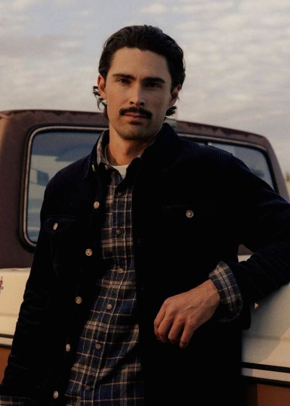 A man wearing a dark corduroy jacket over a blue and brown plaid button-up shirt, leaning against a vintage truck outdoors.