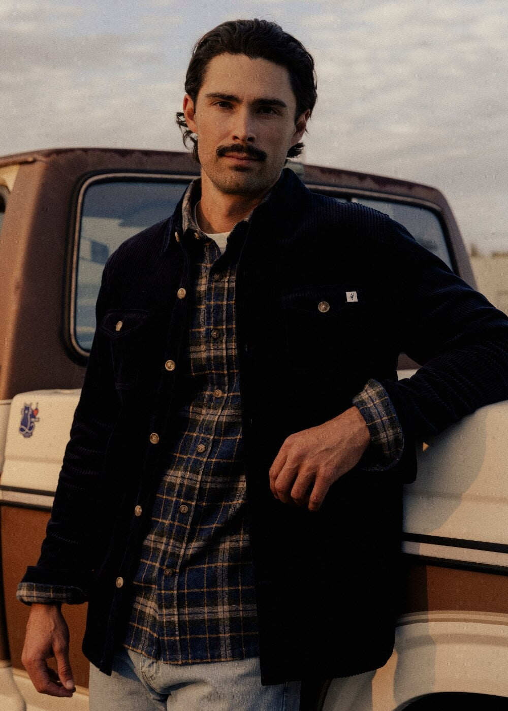 A man wearing a dark corduroy jacket over a blue and brown plaid button-up shirt, leaning against a vintage truck outdoors.