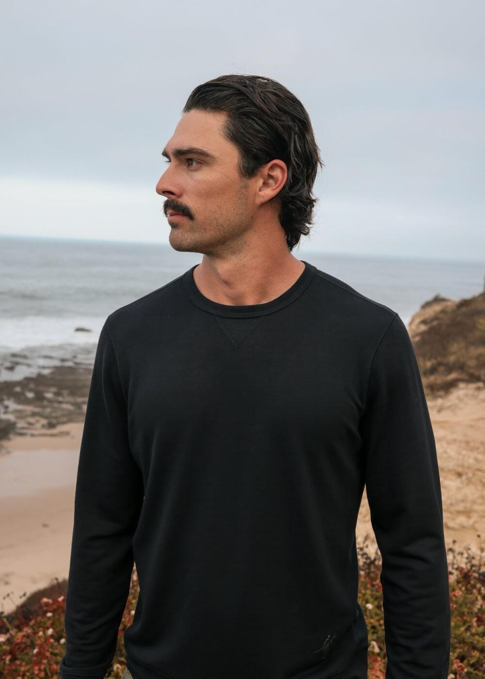 A man with a mustache wearing a black long-sleeve crewneck shirt, standing outdoors on a sandy coastal cliff with the ocean in the background.