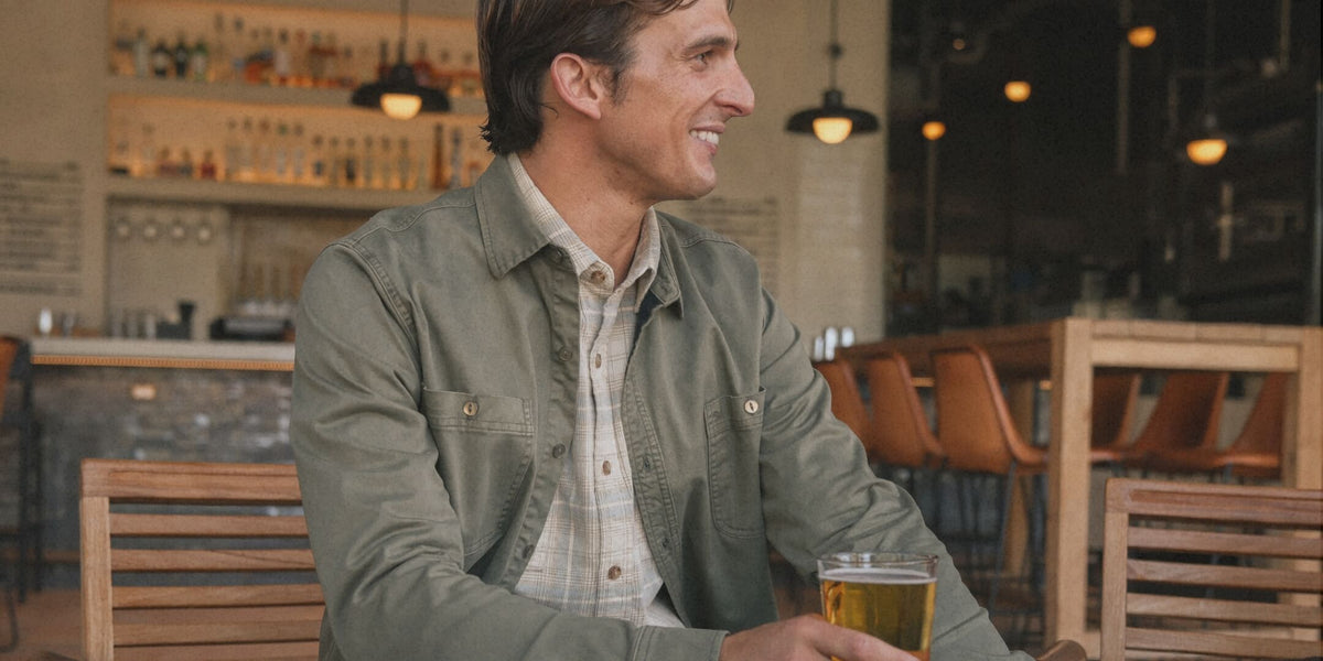 A man sitting at a wooden table in a bar or cafe, wearing an olive green utility overshirt over a light plaid button-down shirt, holding a glass of beer.