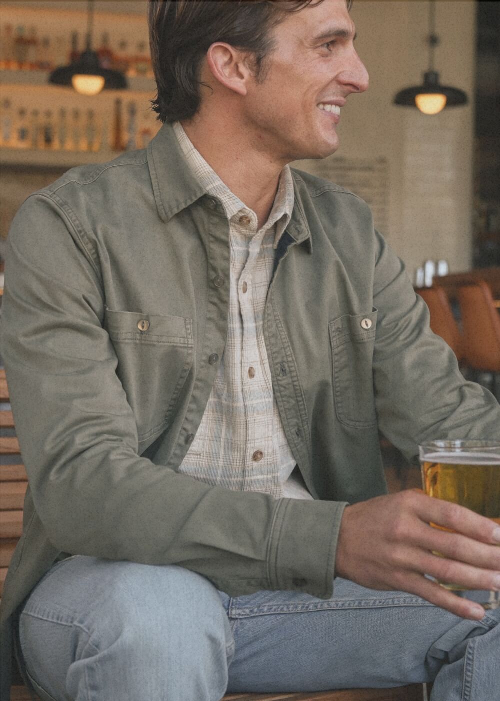 A man wearing an olive green utility overshirt over a beige plaid button-down shirt and light blue jeans, sitting in a bar and holding a glass of beer.