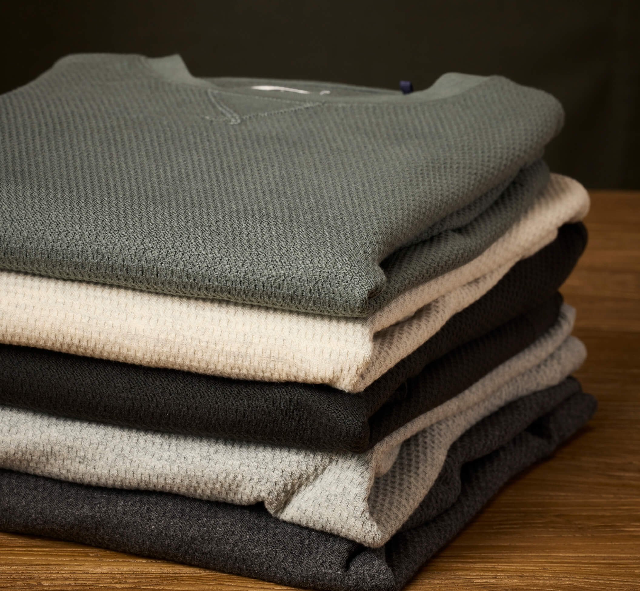 A stack of folded knit crewneck sweaters in muted gray, cream, black, light gray, and charcoal, arranged on a wooden surface against a dark background.