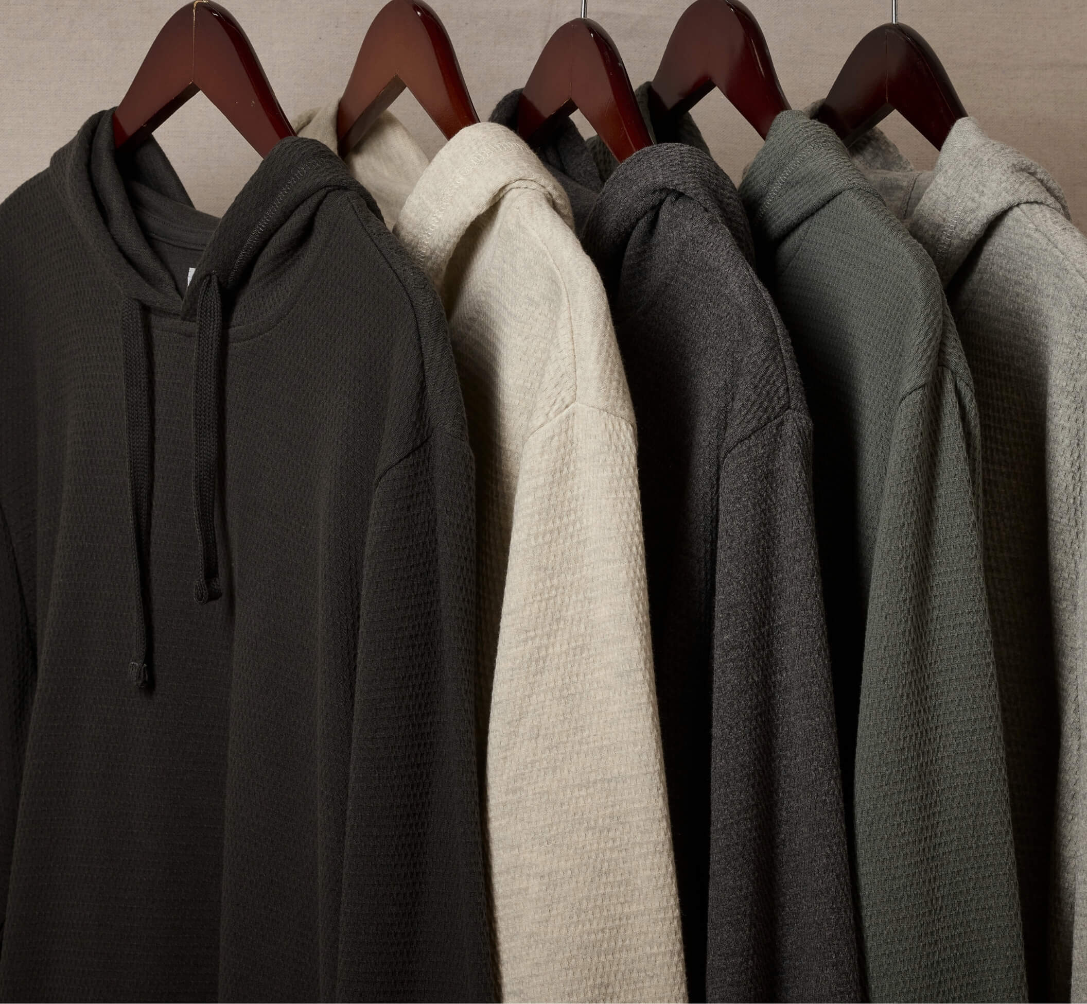 A row of waffle-knit hoodies hanging on wooden hangers against a light beige wall. The hoodies are shown in multiple colors, including charcoal gray, cream, dark gray, sage green, and light gray.