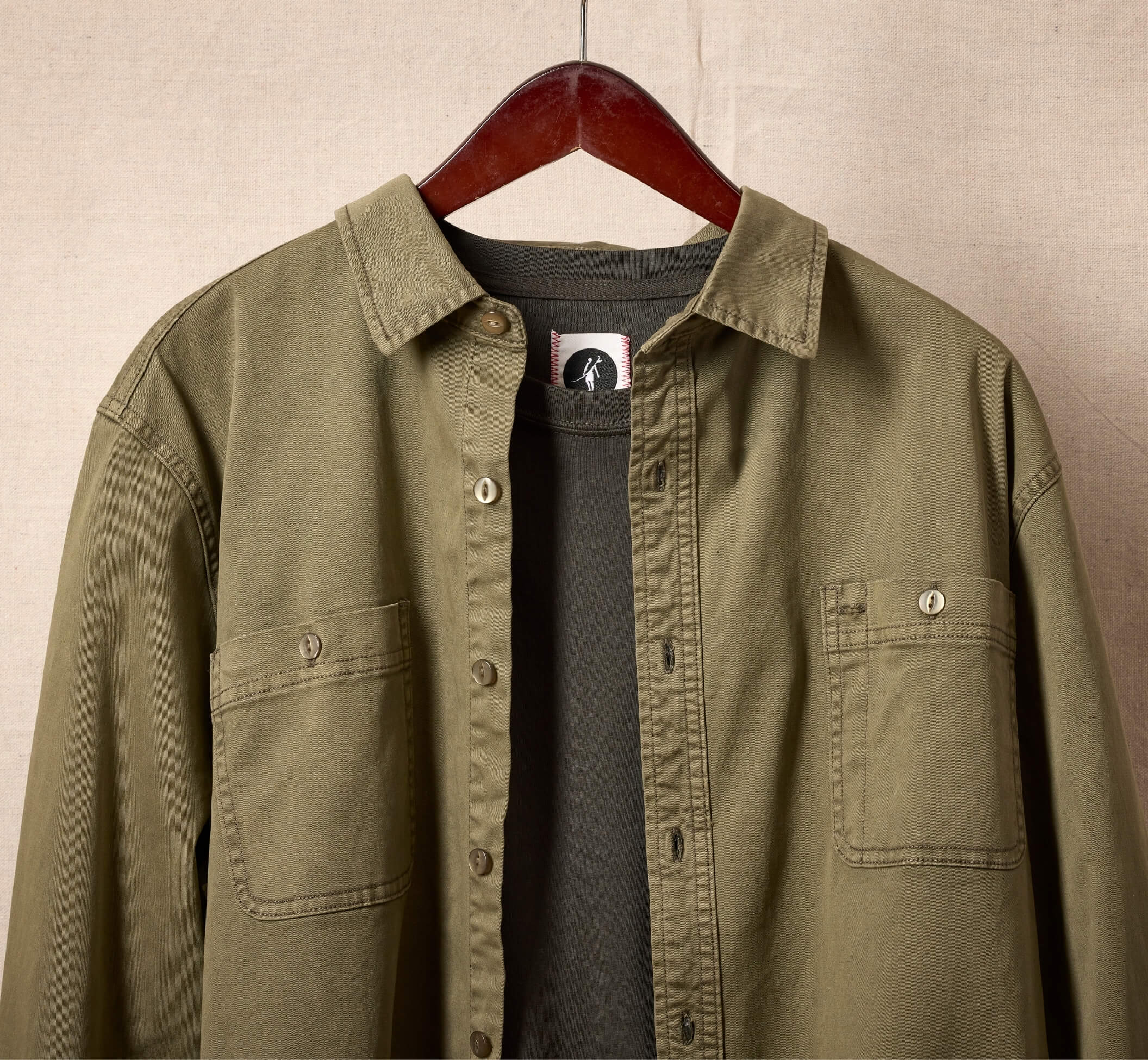 Garment dyed green cotton shirt, layered over a crewneck sweater on a hanger with a canvas studio background.