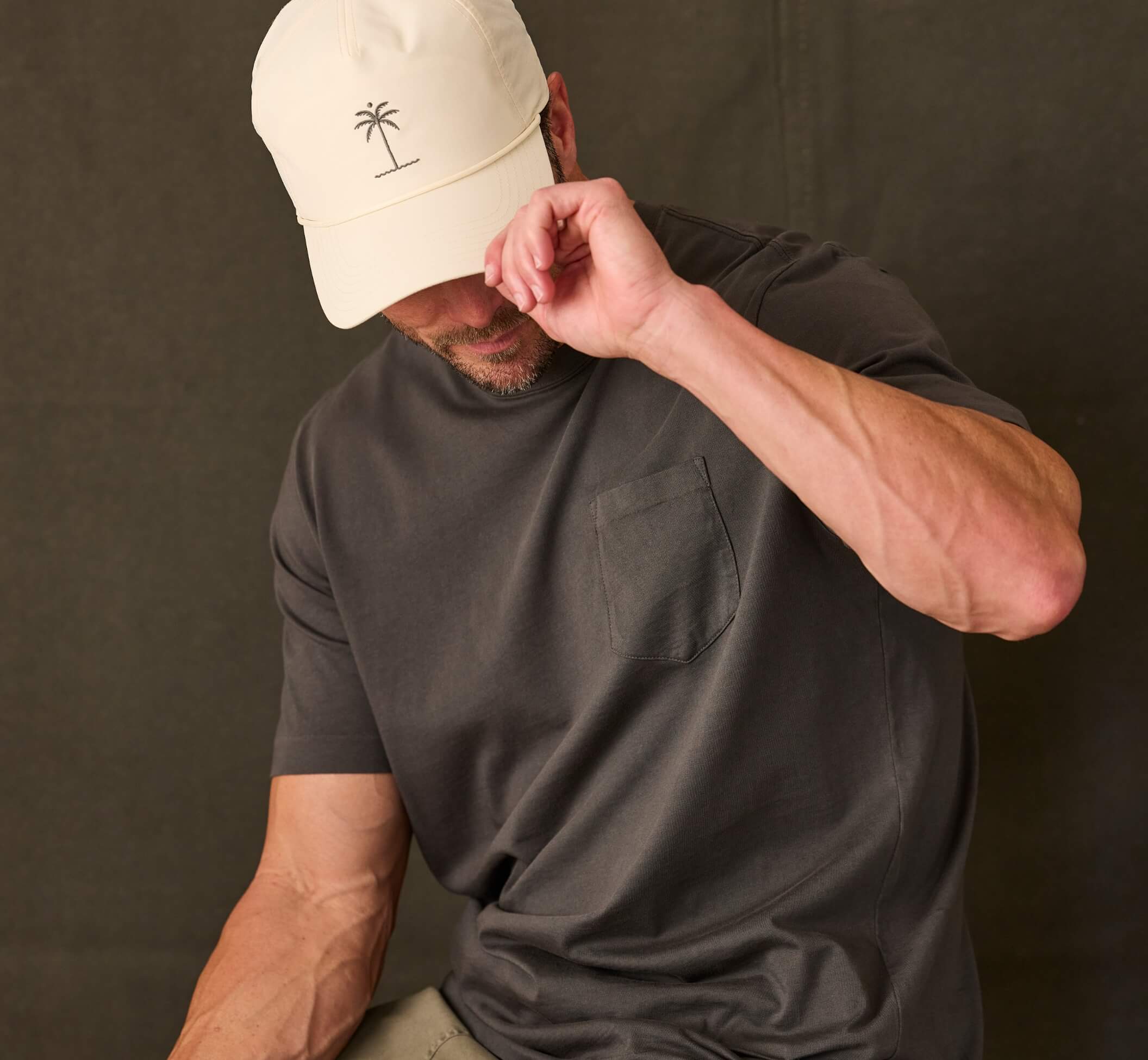 A man wearing a dark charcoal gray short-sleeve pocket t-shirt and a cream-colored baseball cap with a small palm tree logo, posing against a dark gray studio backdrop.