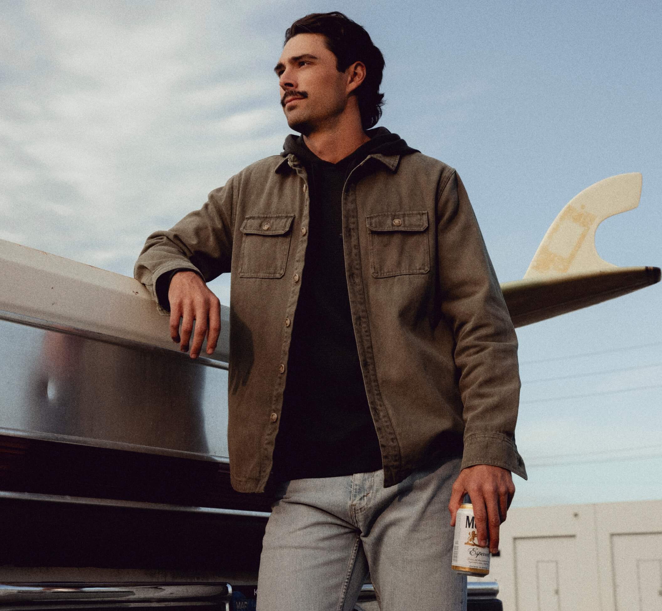 A man wearing a brown overshirt over a black hoodie and light gray jeans, leaning against a boat with a surfboard in the background.