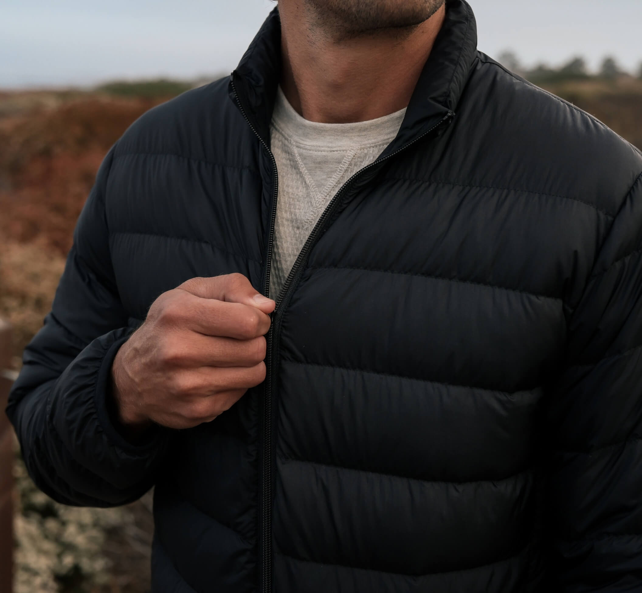 A close-up of a man wearing a black puffer jacket over a light gray t-shirt, with his hand adjusting the zipper, set against an outdoor blurred landscape.