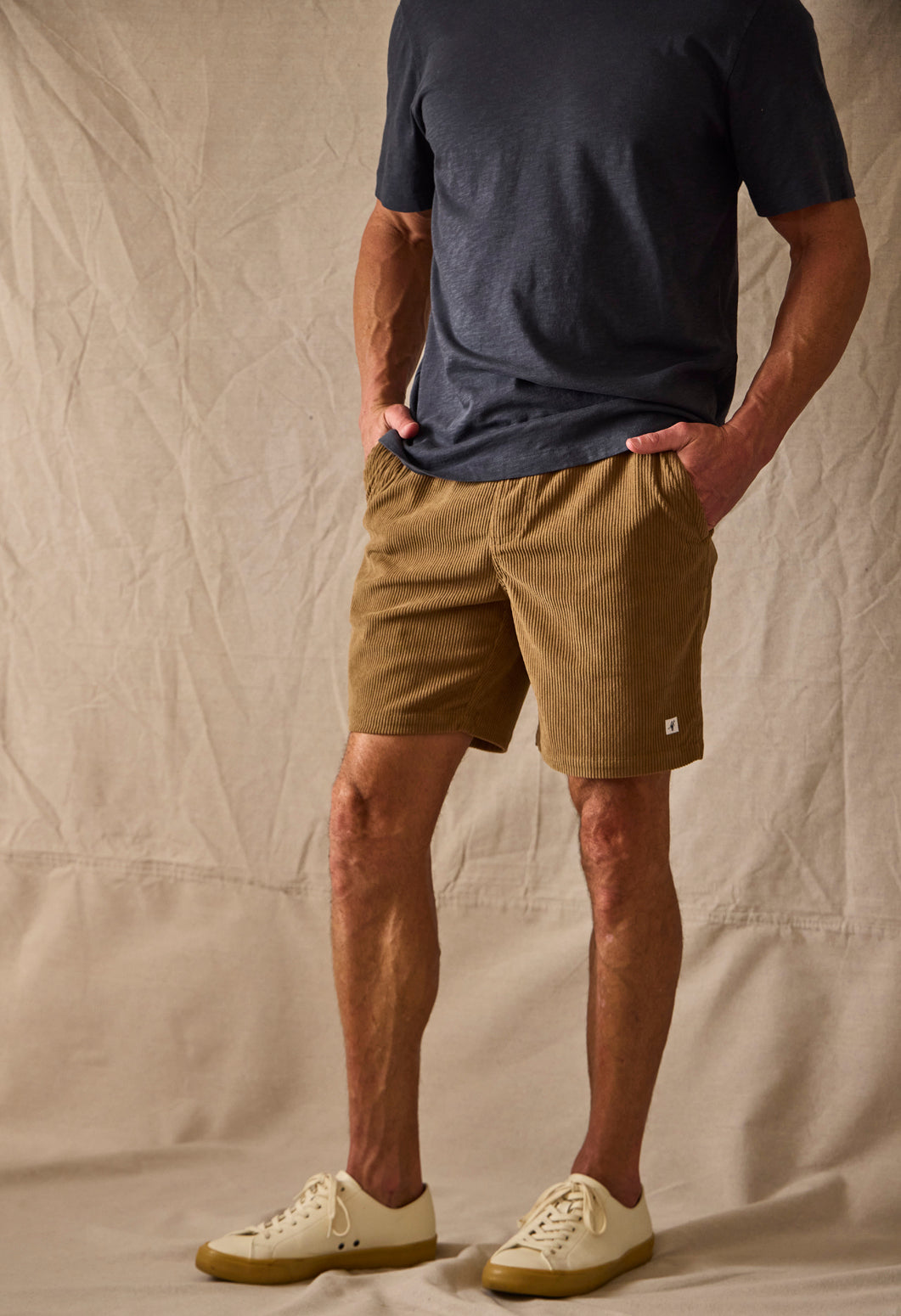 Man wearing brown shorts against a tan background with hand in pocket 