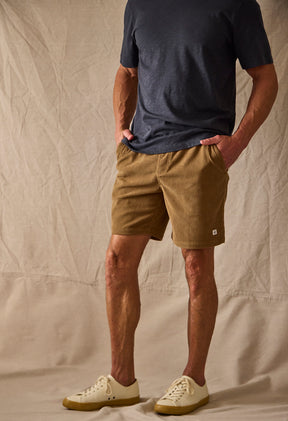 Man wearing brown shorts against a tan background with hand in pocket 