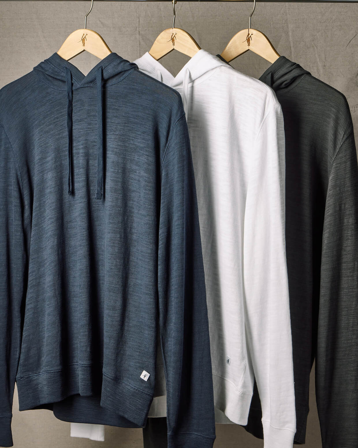 Three hoodies in blue, white, and black hanging on wooden hangers against a gray background.