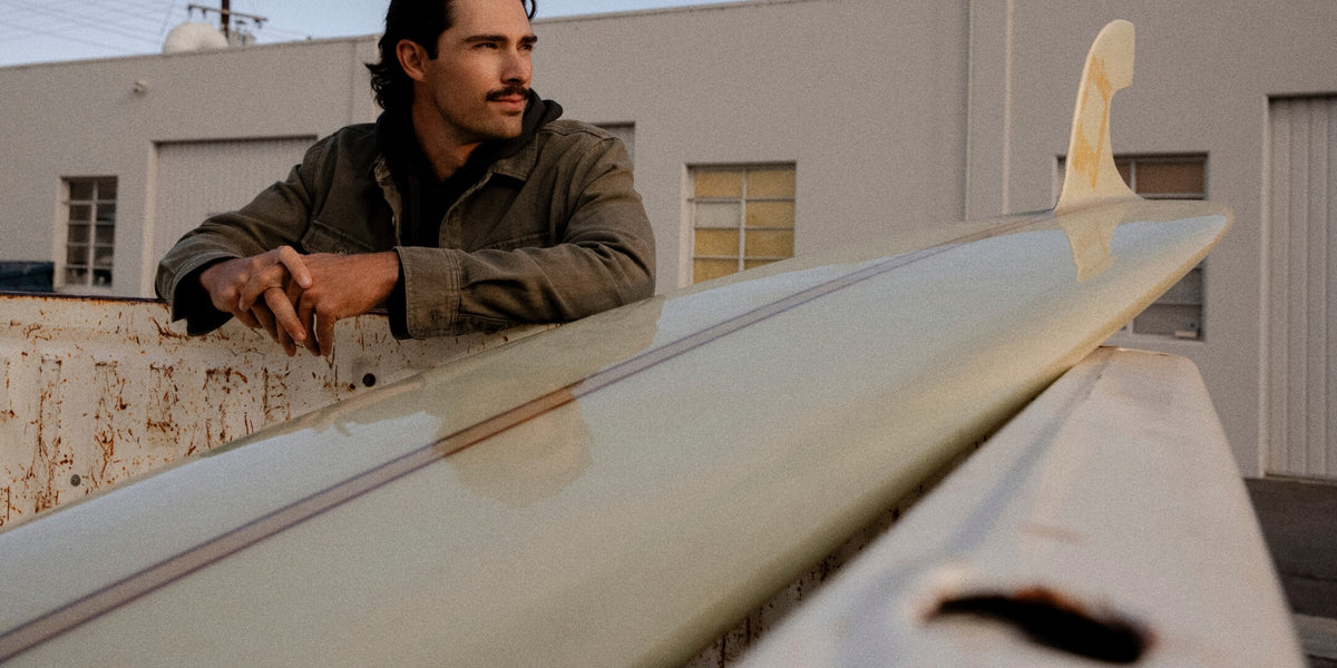 A man in a brown jacket leans over a surfboard in an outdoor industrial setting, with a building and windows in the background.