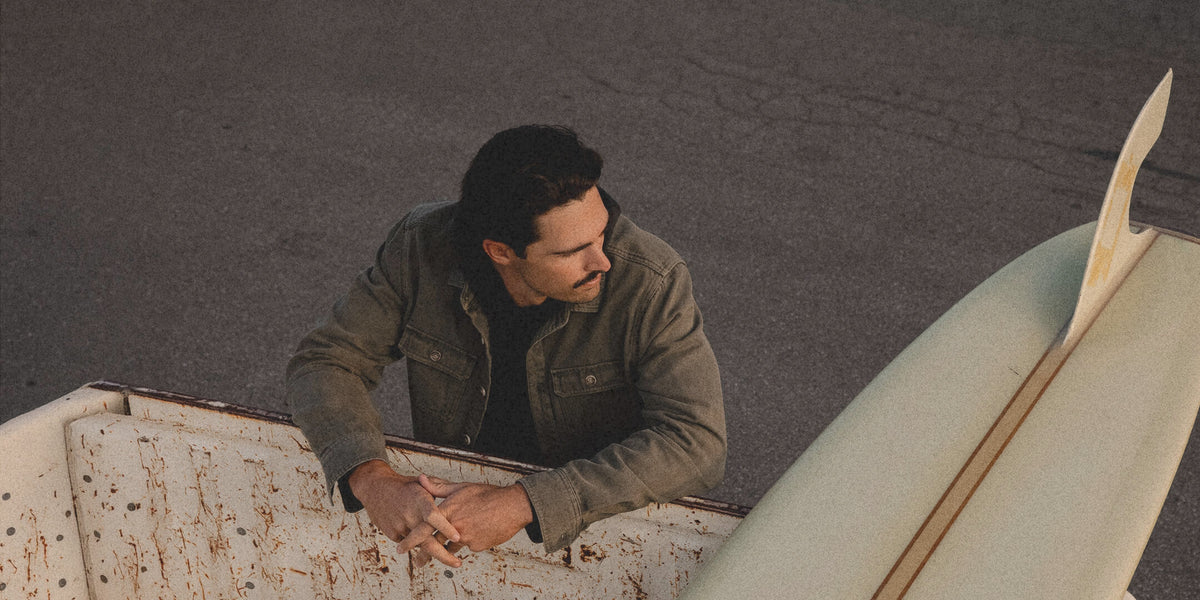 A man in a brown jacket leans over the edge of a rusty white truck bed, looking off-camera next to a surfboard on an asphalt road.