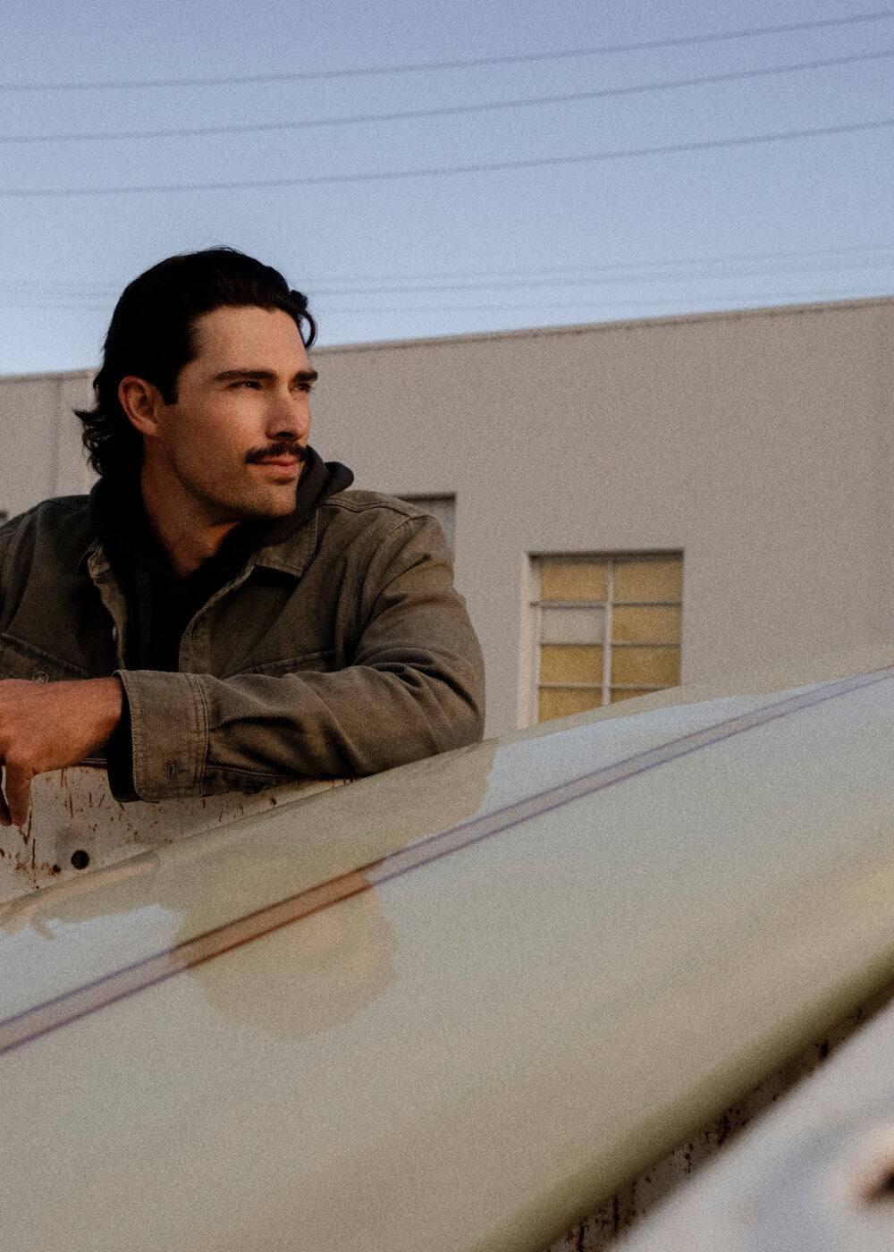 A man with dark hair and a mustache leans on a car outdoors, wearing a brownish-gray jacket. The background shows a light-colored building under a pale blue sky.