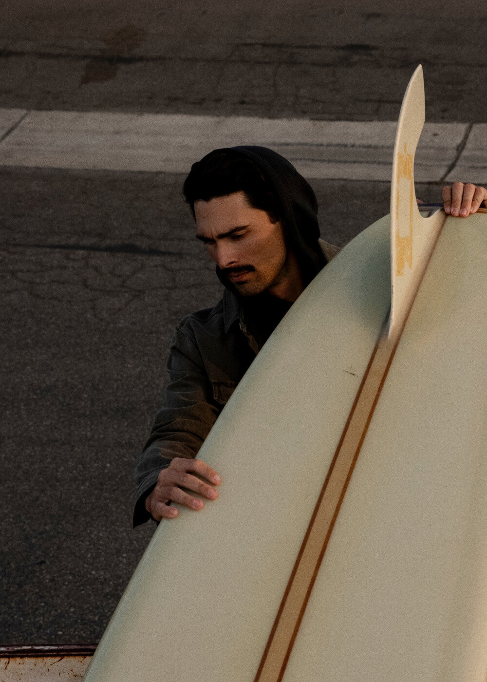A man in a dark hooded jacket holds a large cream-colored surfboard on an asphalt surface at sunset.