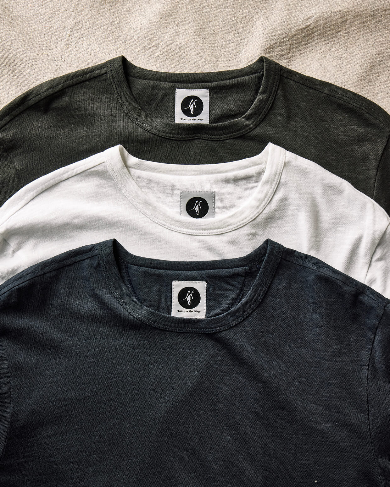 Three t-shirts in black, white, and gray with a brand logo on a beige fabric background. 