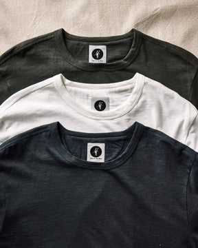 Three t-shirts in black, white, and gray with a brand logo on a beige fabric background. 